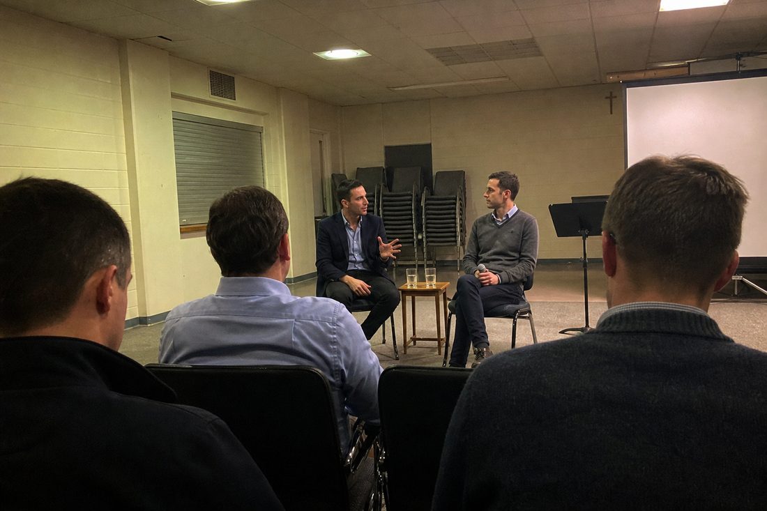 Illustration of the October evening: two speakers seated in conversation at the front of a parish hall, seen from within the audience.