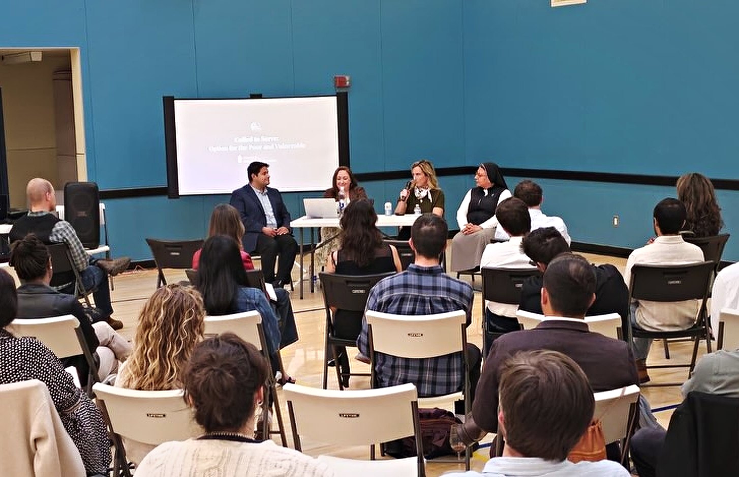 A past evening seen from the back of the parish hall: a full room of seated people facing a four-person panel at a table under a projected slide.