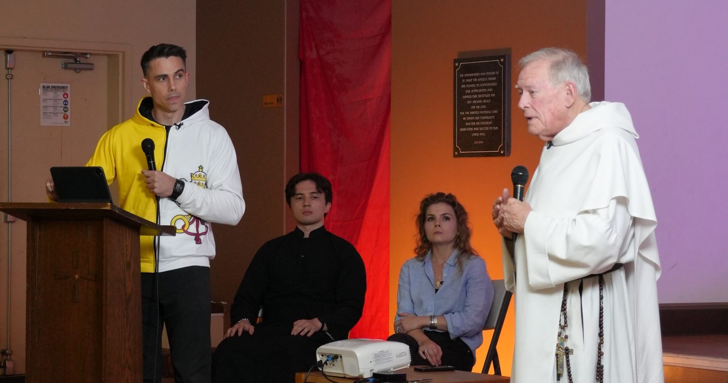 Fr. Michael Sweeney, OP speaking into a handheld microphone in his white Dominican habit while Sean Bryan stands at the lectern, a deep red curtain behind them.