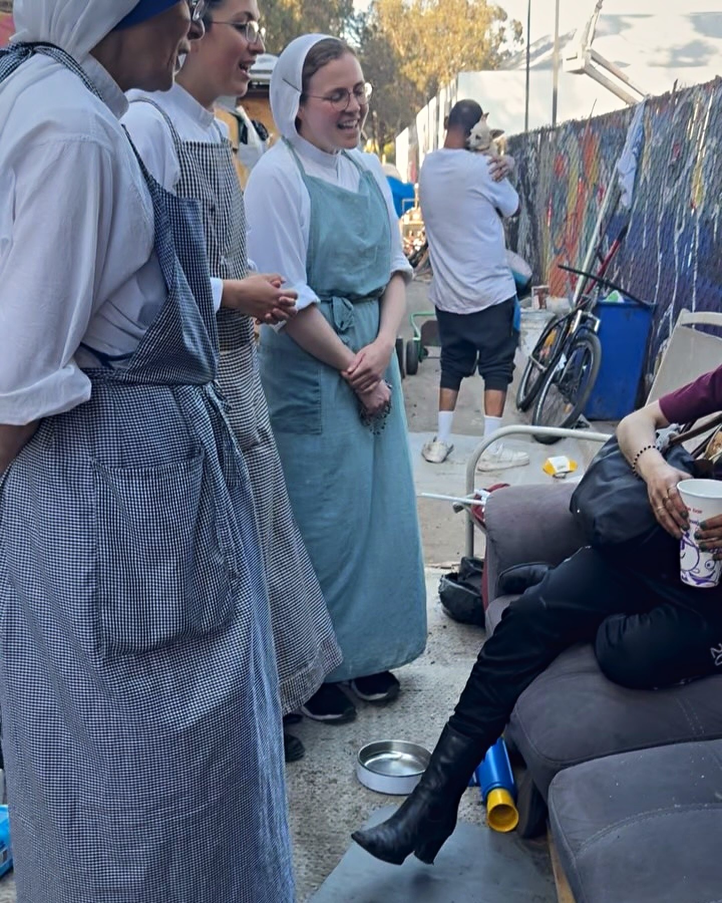 Three religious sisters in aprons standing and laughing with a person seated on a couch at a street encampment, a bicycle and a painted fence behind them.